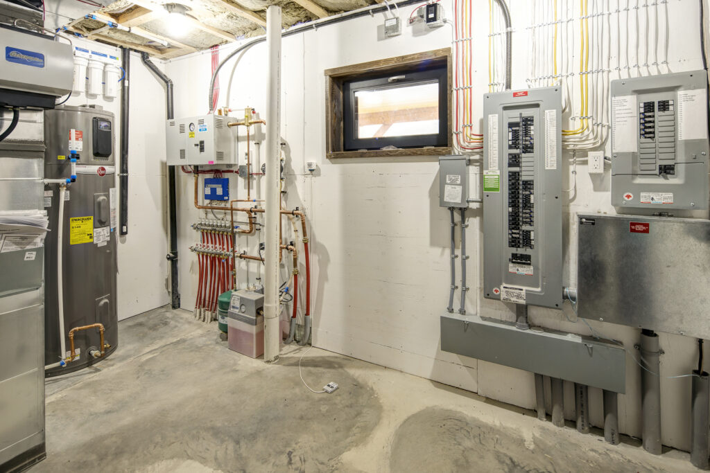 Basement utility room with exposed pipes, electrical panels, water heater, and a small window. Concrete floor and unfinished ceiling are visible—an essential space in many energy efficient homes.