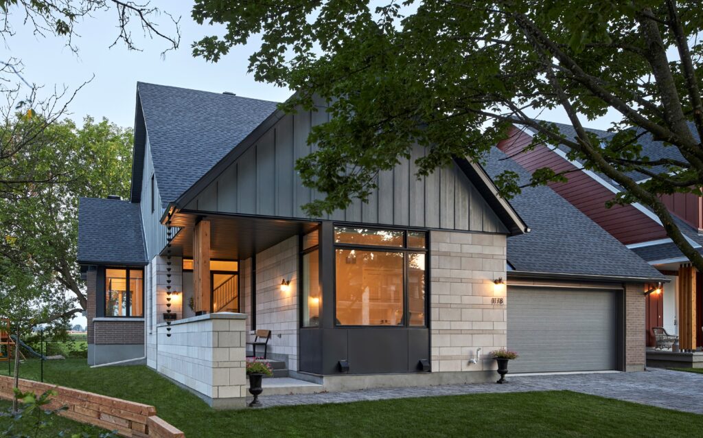 Modern net zero home with a gray roof, large front windows, and a double garage. Warm lights illuminate the entryway. Trees and grass surround the energy efficient house. A wooden bench is on the porch, and potted plants sit by the entrance.