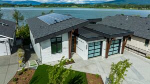Modern lakefront house with large windows, a black roof featuring solar panels, two glass garage doors, landscaped yard, and a water view with green hills and cloudy skies in the background—perfect for Home design inspiration or anyone seeking a Renovated Net Zero lifestyle.