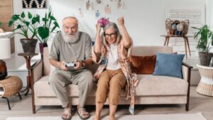 An older man and woman sit on a couch; he plays a video game with a controller, while she cheers, raising her arms in excitement. The cozy room—decorated with plants and personal touches—reflects their commitment to creating a home for life, making aging in place both joyful and vibrant.