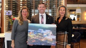 Three people in business attire stand indoors, smiling and holding a large rendering of a modern building with parking lots—a project by Solmar Development Corporation. Wine bottles are displayed on a wall behind them, emphasizing an elegant setting as they celebrate giving back to the community in support of the St. Catharines hospital.