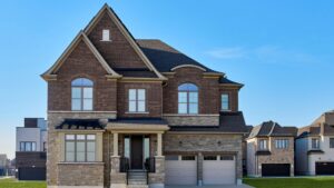 A large two-story house with a brick and stone exterior, arched windows, a covered front porch, and a double garage sits under a clear blue sky in a suburban neighborhood—an exceptional example of award winning net zero homes that blend elegant design with outstanding energy efficiency.