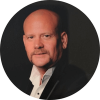 A bald man with a mustache, wearing a dark suit jacket and white shirt, poses for a formal portrait against a dark background.