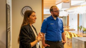Two people stand in a brightly lit office space. The woman on the left is smiling and speaking about giving back, while the man beside her in a blue shirt listens. A clock and modern dining area are visible in the background.