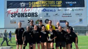 A soccer team poses with a trophy in front of a Constructors Cup Corporate Soccer Tournament banner outdoors, smiling and dressed in matching black shirts, proudly representing the London Community Foundation.