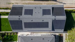 Aerial view of a house roof with multiple rectangular solar panels installed, surrounded by green lawns and a fence, highlighting a modern approach to renewable energy and net zero performance homes.