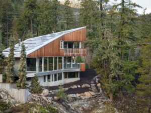 Modern Net Zero home with wood and glass exterior sits among tall pine trees on a rocky hillside. The home features large windows and sloped roofs, blending seamlessly with the natural forest surroundings while exemplifying sustainable net zero home building.