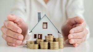 A person’s hands encircle a small model house with stacked coins in front of it, symbolizing home investment, savings, or protection of property and finances—perfect for illustrating the benefits of home renovations or renovation tax credits.