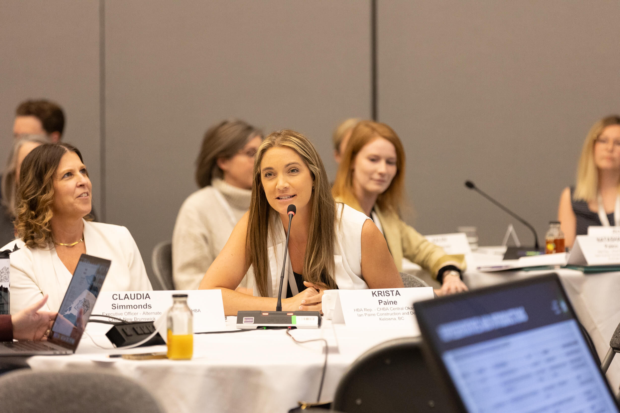 A woman speaks into a microphone at a conference table, surrounded by others. Name cards in front of her read Krista Paine and Claudia Simmonds. Laptops, papers, and drinks are visible on the table.