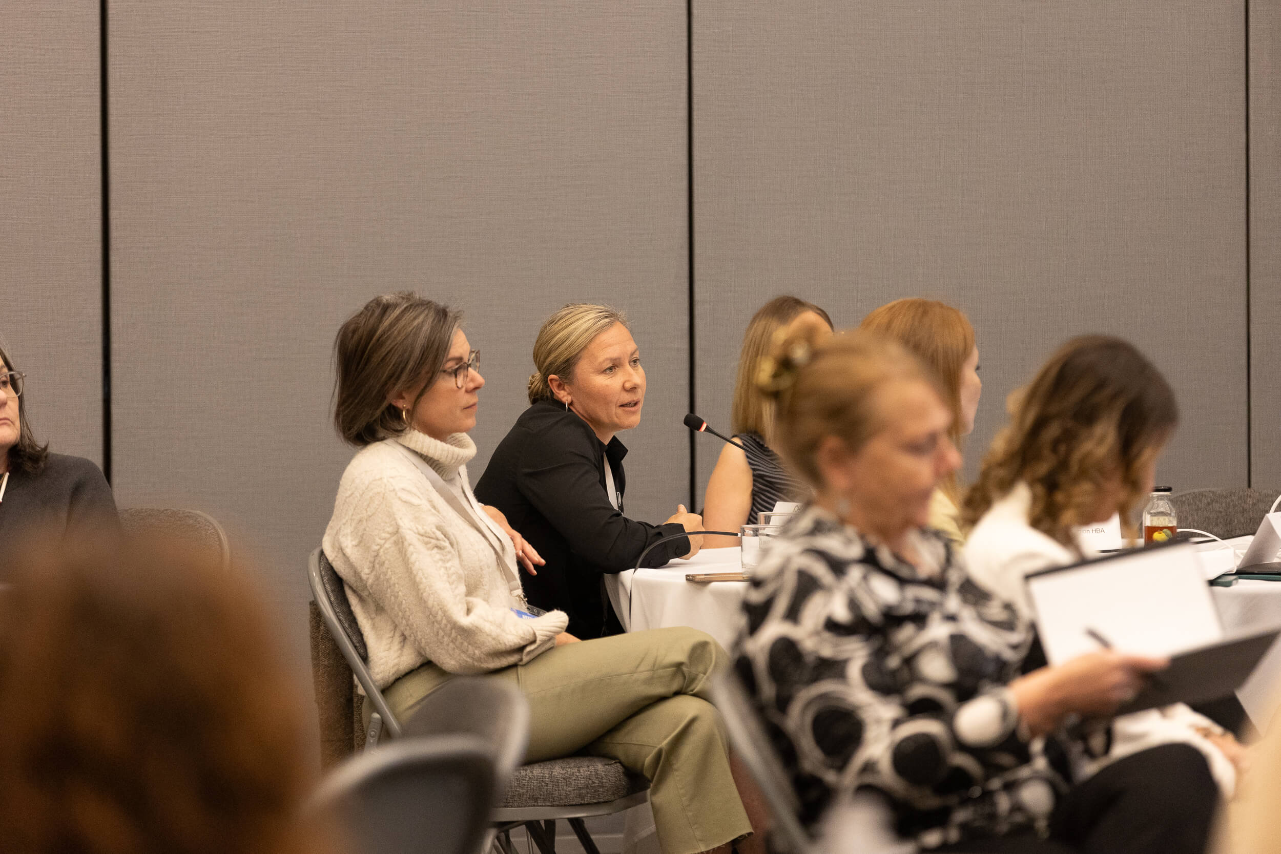 A woman speaks into a microphone while seated at a table among other attendees during an indoor conference or meeting. People around her listen or take notes.