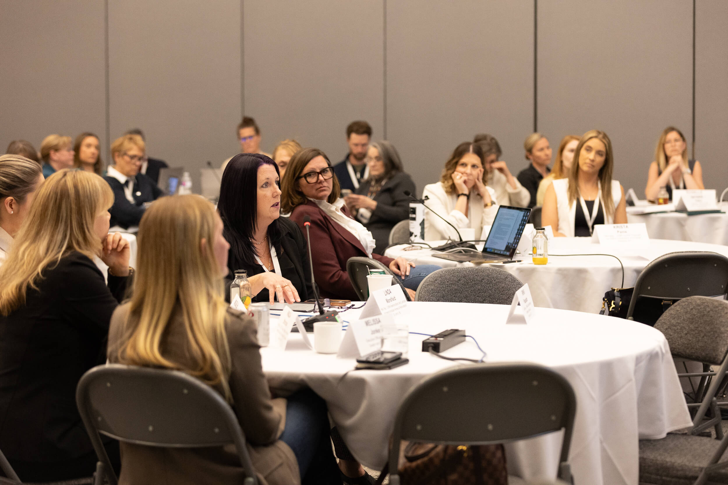 A group of people, mostly women, sit around round tables in a conference room, attentively listening to a speaker. Some have laptops and notepads; name tags and bottled drinks are visible on the tables.