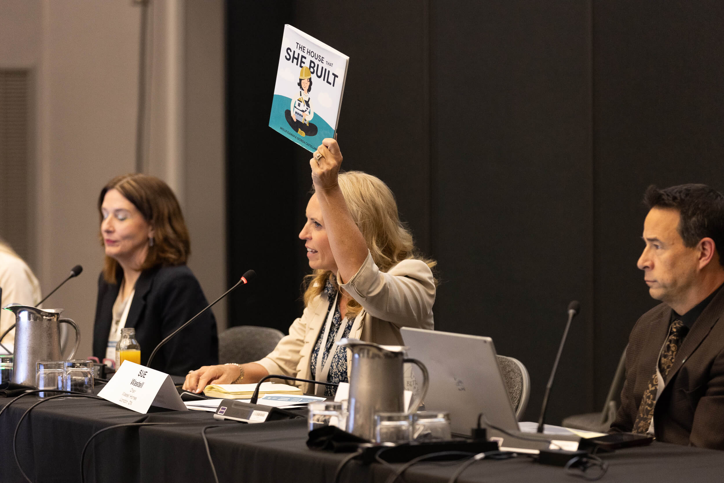 A woman sitting at a conference table holds up a book titled The House That She Built while speaking into a microphone; two other panelists sit beside her.