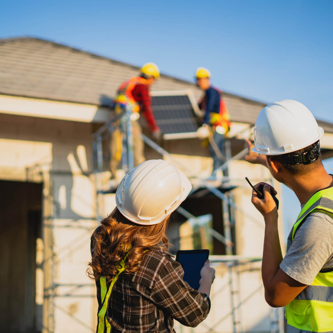 Two construction workers install a solar panel on a house roof, while two supervisors wearing hard hats and safety vests observe and communicate via radio and tablet in the foreground.