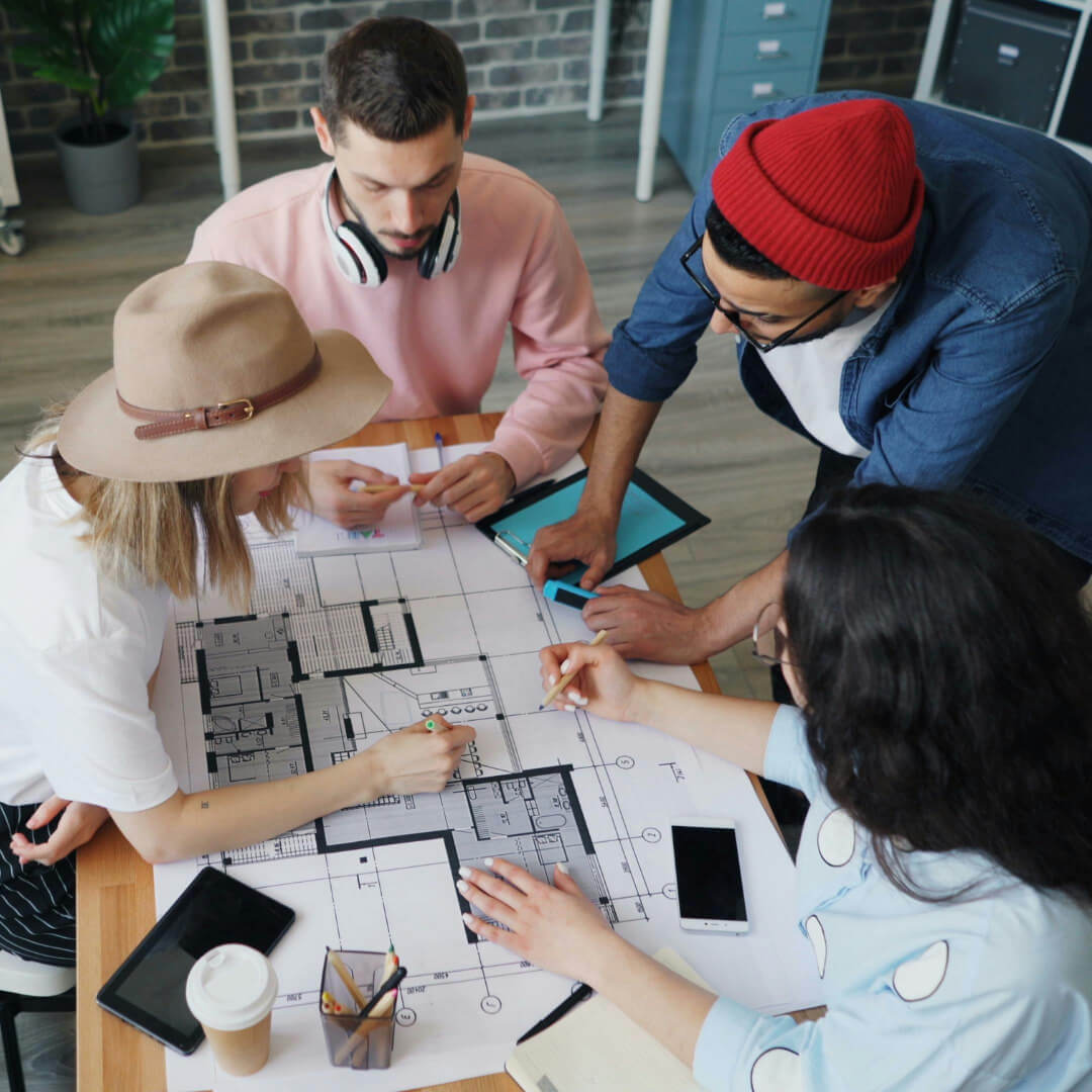 Four people are gathered around a table, actively collaborating on a floor plan drawing. They are discussing and pointing at the blueprint, with smartphones, notebooks, and stationery scattered on the table.