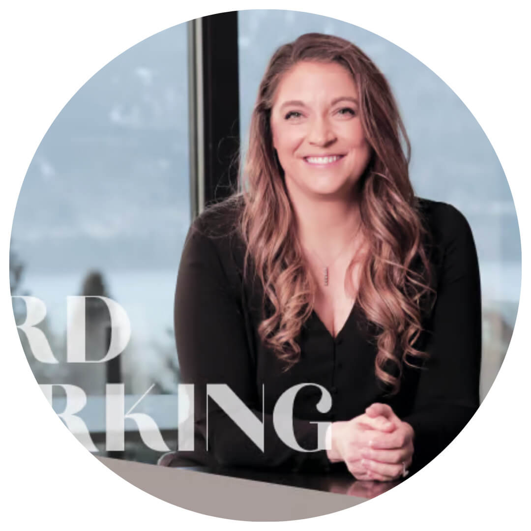 A woman with long wavy hair, wearing a black top, sits smiling at a table with her hands clasped. There is a window with a scenic outdoor view behind her. Partial text overlays the image.