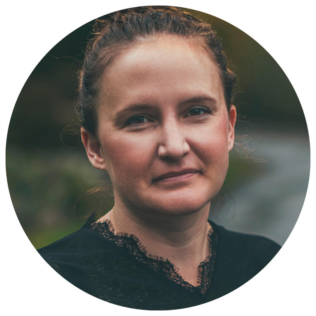 A woman with light skin and brown hair pulled back looks at the camera with a neutral expression, wearing a dark top and standing outdoors with a blurred natural background.