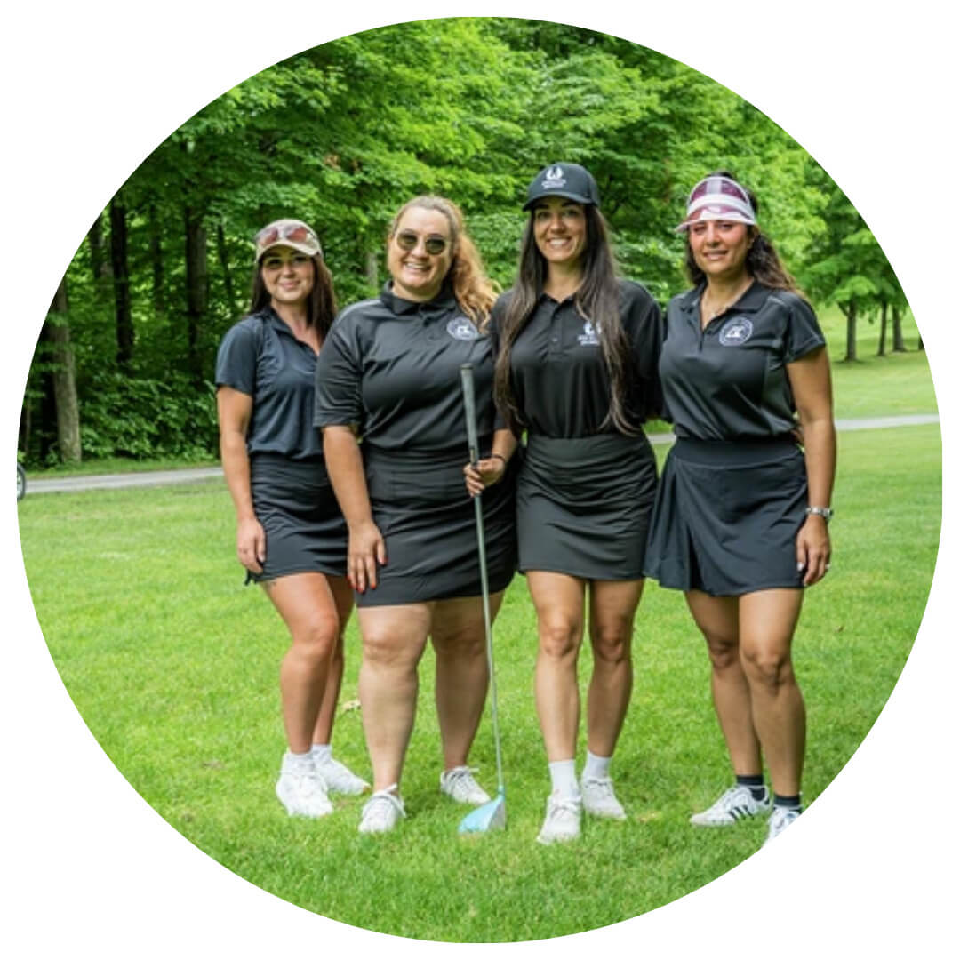 Four women in matching black golf outfits and hats stand together on a green lawn with trees in the background, smiling for the camera. One woman holds a golf club.