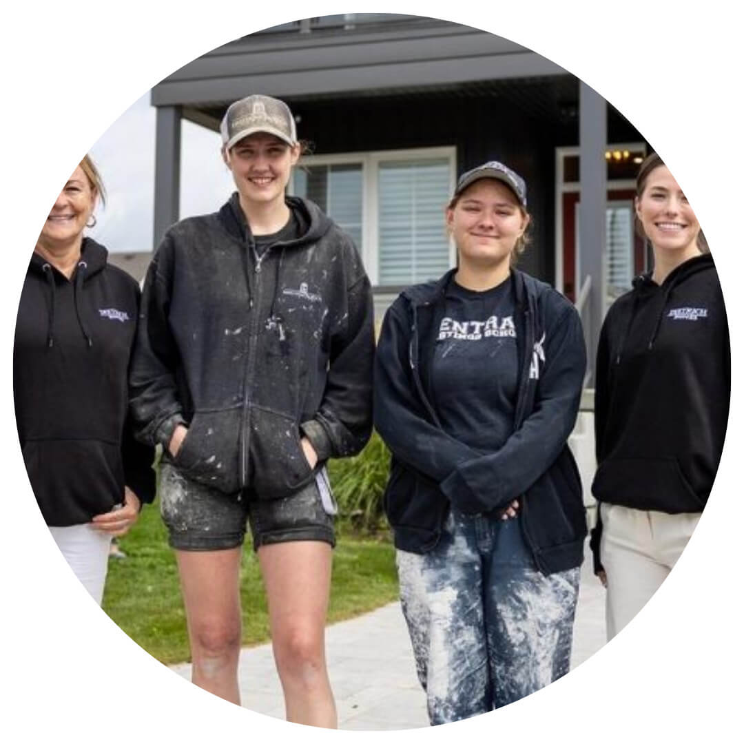 Four people stand outdoors in front of a house, smiling. Two wear dark hoodies and pants with paint stains, suggesting painting or construction work. The group appears relaxed and happy.