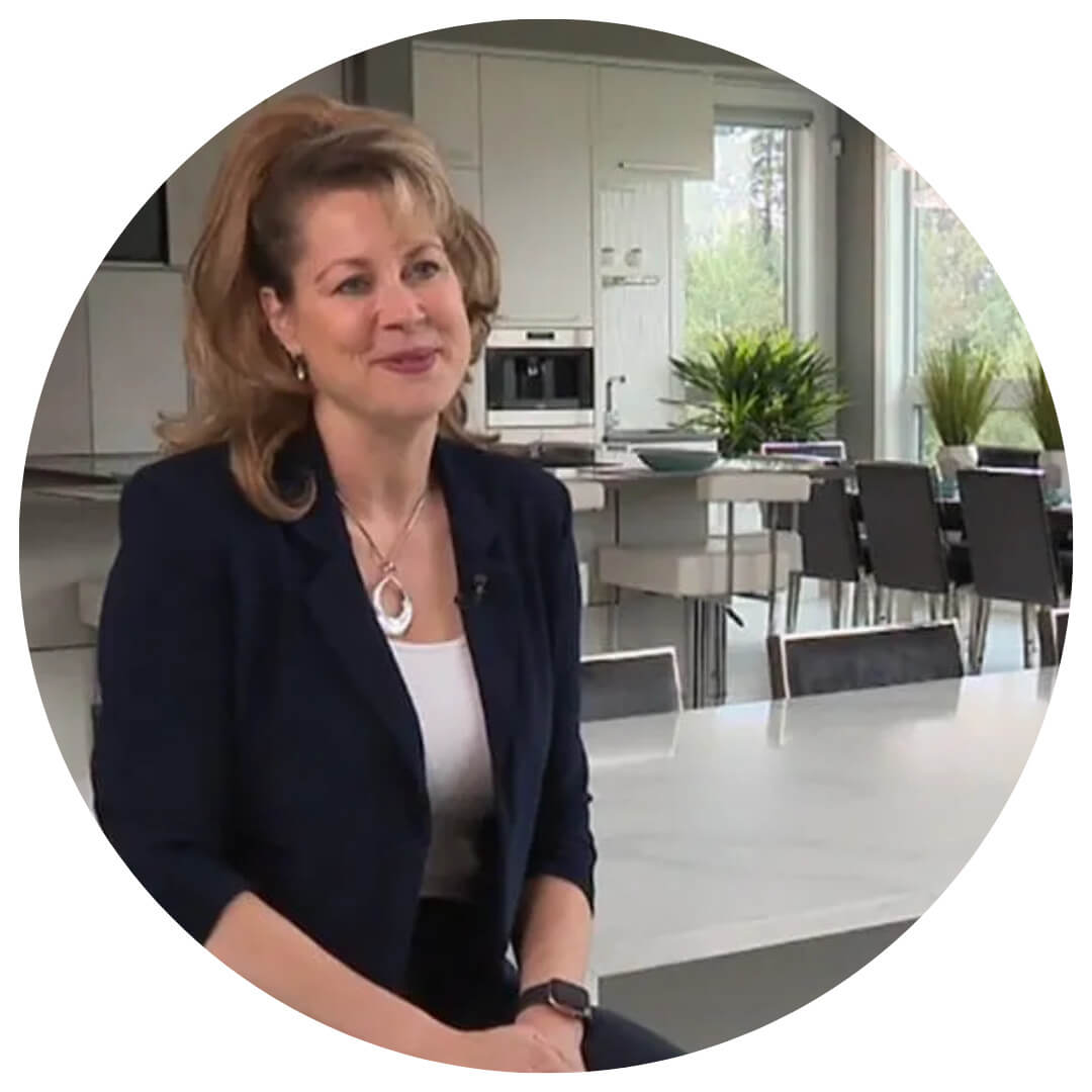 A woman with light brown hair sits at a white countertop in a modern kitchen, smiling. She wears a navy blazer, white top, and necklace. Green plants and large windows are visible in the background.