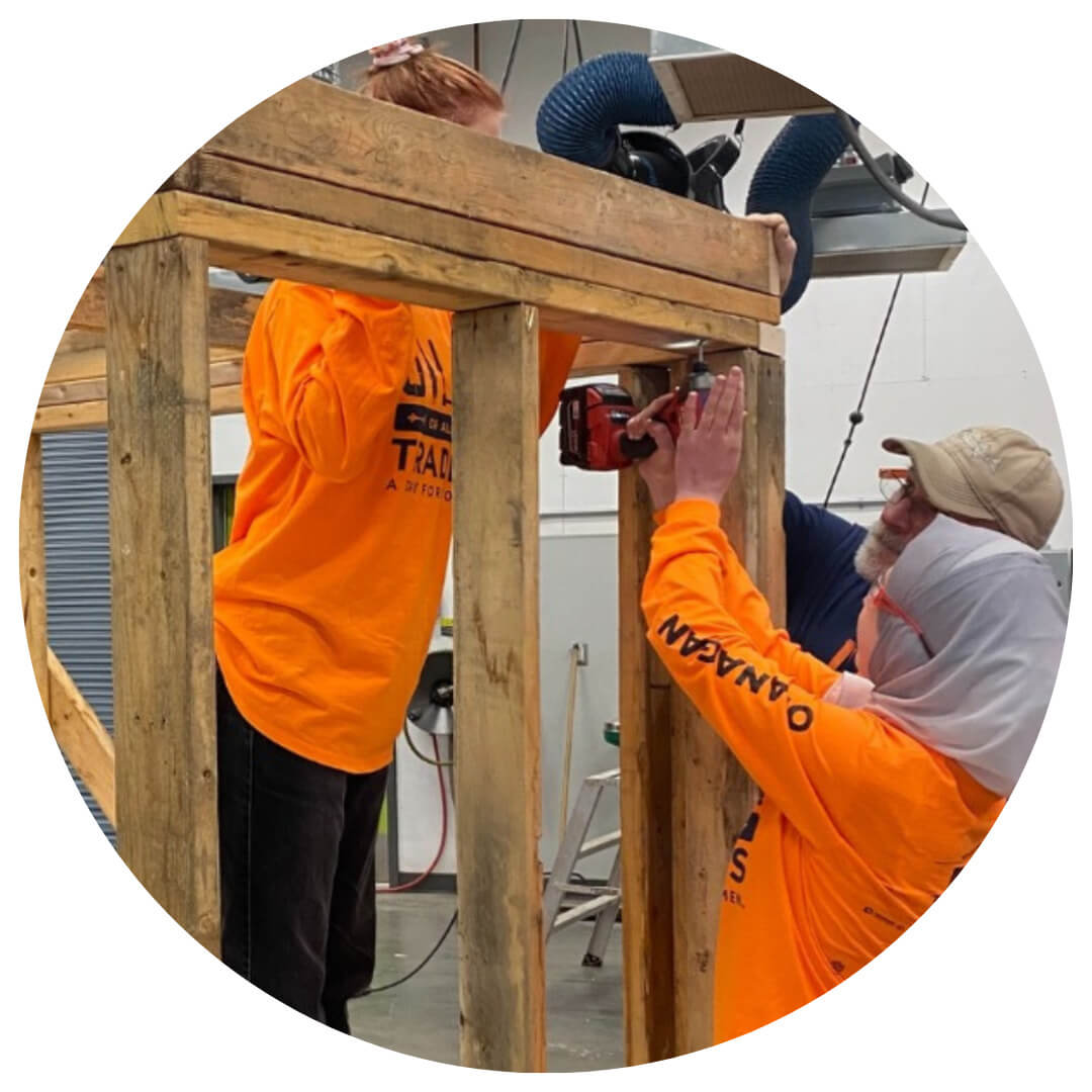 Two people in bright orange shirts use a power drill to build a wooden structure indoors. One person stands on the left, while the other on the right wears a hat and looks up, focusing on their task.
