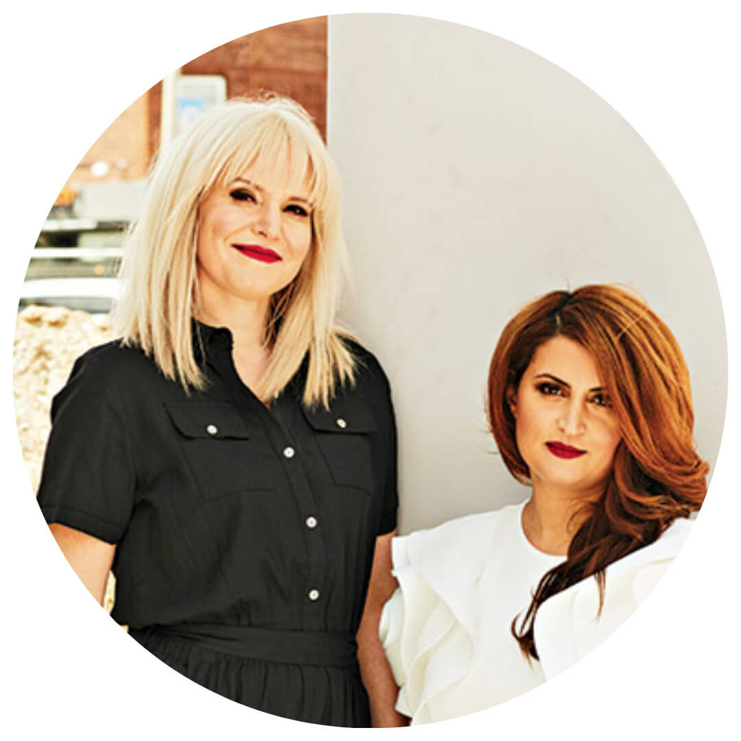 Two women pose together. One has straight, blonde hair and wears a black dress; the other has long, wavy, brown hair and wears a white ruffled top. Both are smiling and standing by a light-colored pillar.