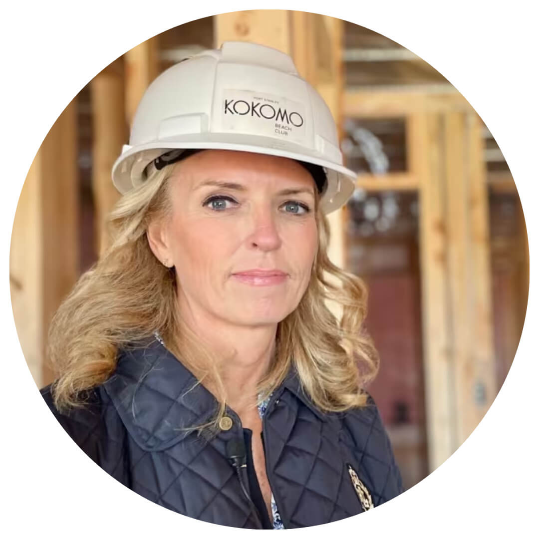 A woman with blonde hair wearing a white hard hat labeled KOKOMO and a navy quilted jacket stands in a building under construction with exposed wooden beams in the background.