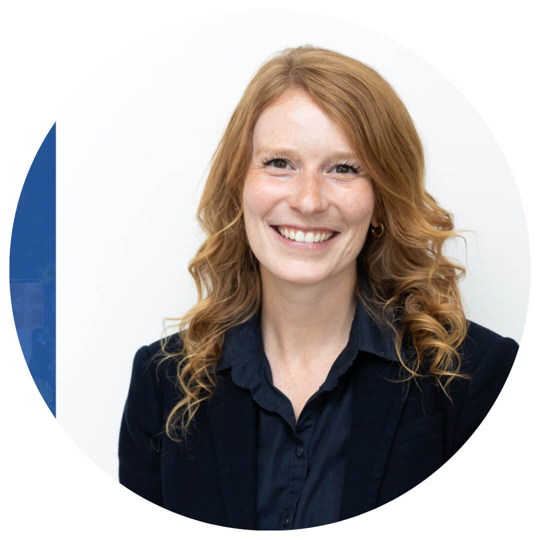 Smiling woman with wavy red hair wearing a dark blazer and navy blouse, standing against a plain white background.