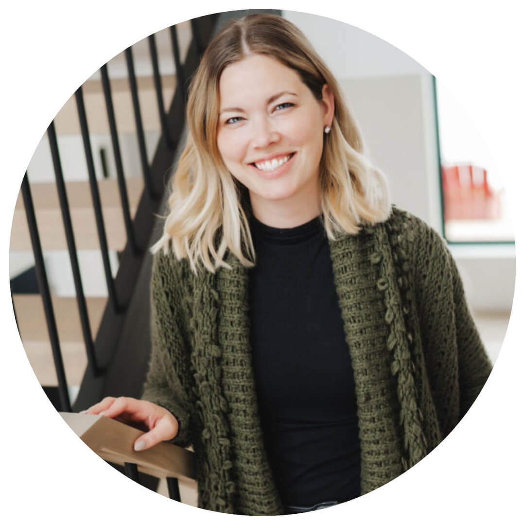 A smiling woman with blonde hair wearing a green knit cardigan and black top stands indoors by a staircase, looking at the camera.