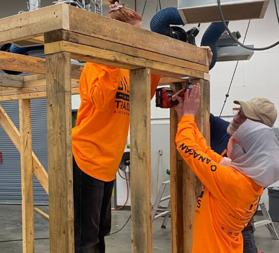Two people in bright orange shirts assemble a wooden structure indoors. One holds a power drill while the other supports the wood, both focused on their work. Industrial equipment and tools are visible in the background.