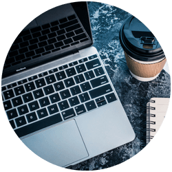 A close-up of a laptop keyboard, a spiral notebook, and a takeaway coffee cup on a dark marble surface.