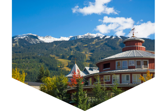 A round wooden building with a weather vane stands against green pine trees and snow-capped mountains under a blue sky with scattered clouds, evoking the scenic locations often chosen for CHBA events for the home building industry.