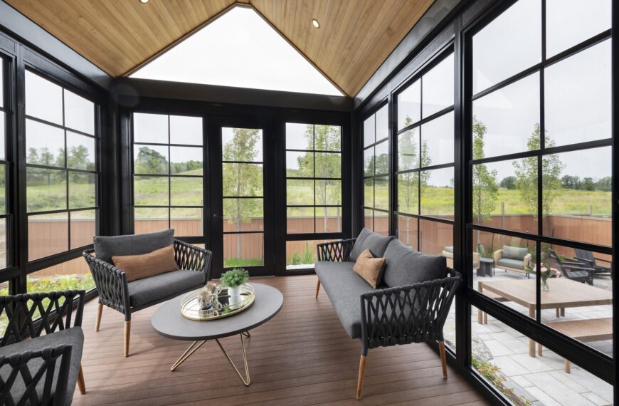 A modern sunroom with large black-framed windows, wooden ceiling, and brown flooring. Two gray sofas and a round coffee table create a bright, airy space in this net zero home overlooking a patio and green yard.