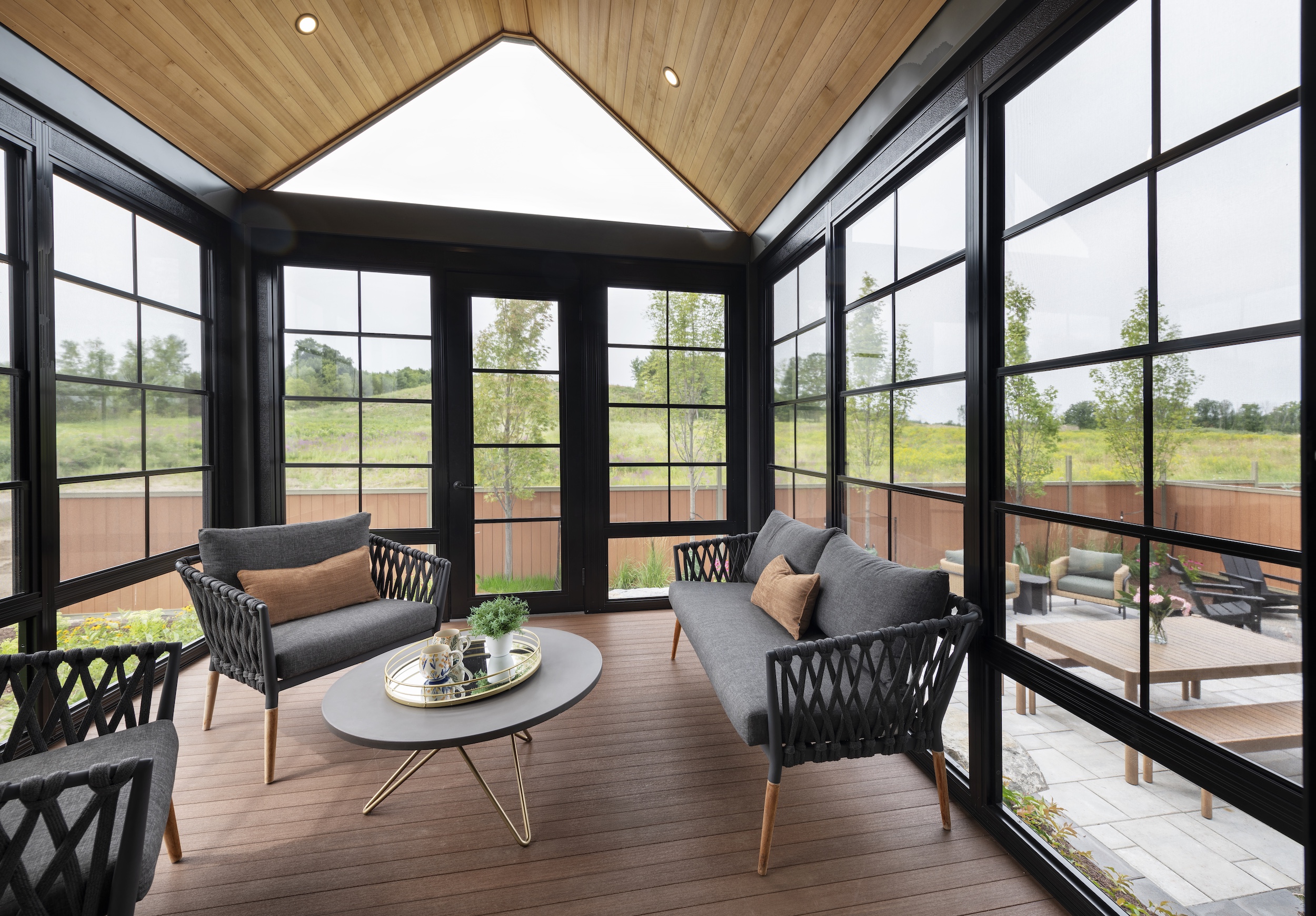 A modern sunroom with large black-framed windows, wooden ceiling, and brown flooring. Two gray sofas and a round coffee table create a bright, airy space in this net zero home overlooking a patio and green yard.