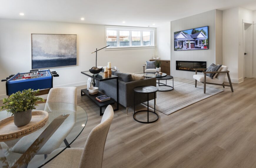 Modern living room with light wood flooring, gray sofa, round glass dining table, armchairs, and foosball table. A wall-mounted TV sits above the fireplace beneath large abstract art. This bright space by Land Ark Homes features recessed lighting.