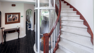 A modern home interior with a curved staircase, a glass elevator, dark wood floors, a small table with a clock and cane, wall art, and an arched entry leading to a bright living area.