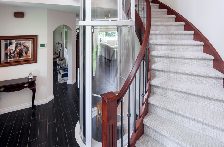 A modern home interior with a curved staircase, a glass elevator, dark wood floors, a small table with a clock and cane, wall art, and an arched entry leading to a bright living area.