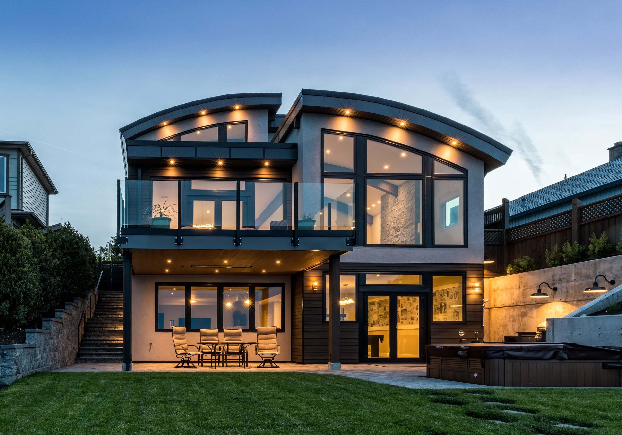 Modern two-story house with large glass windows, curved roof, balcony, covered patio with dining table, outdoor lighting, hot tub, and landscaped backyard at dusk.