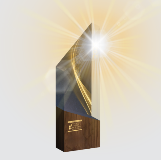 A golden, geometric-shaped award trophy with a bright light shining from its top right corner. The base is made of dark wood with a gold plaque, and the background is softly illuminated.