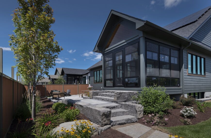 A modern Production Net Zero Home by Land Ark Homes features a screened porch, stone steps, and a landscaped garden with vibrant flowers, shrubs, and a young tree. Enjoy outdoor seating on the stone patio near a wooden fence under the blue sky.