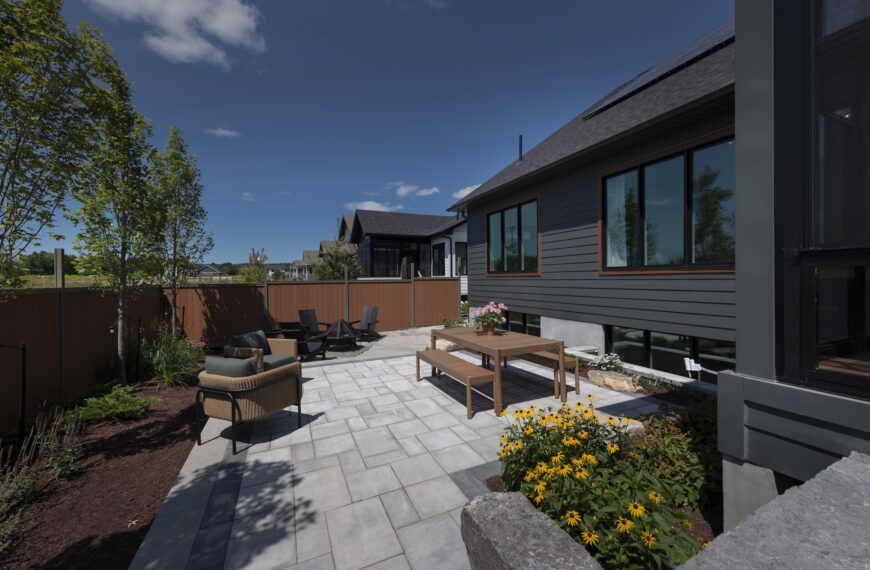 A sunny backyard patio at a Land Ark Homes net zero home features stone pavers, a wooden dining table, yellow flowers in bloom, and an outdoor seating area with a fire pit, all surrounded by a wooden fence and lush trees.