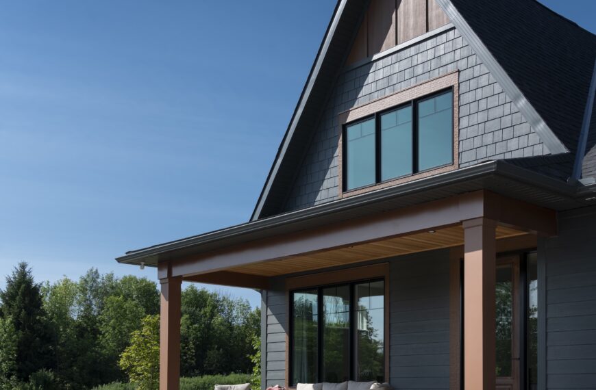 A modern Production Net Zero Home with dark gray siding and large windows features a covered porch with outdoor seating, surrounded by greenery and a landscaped garden under a clear blue sky.