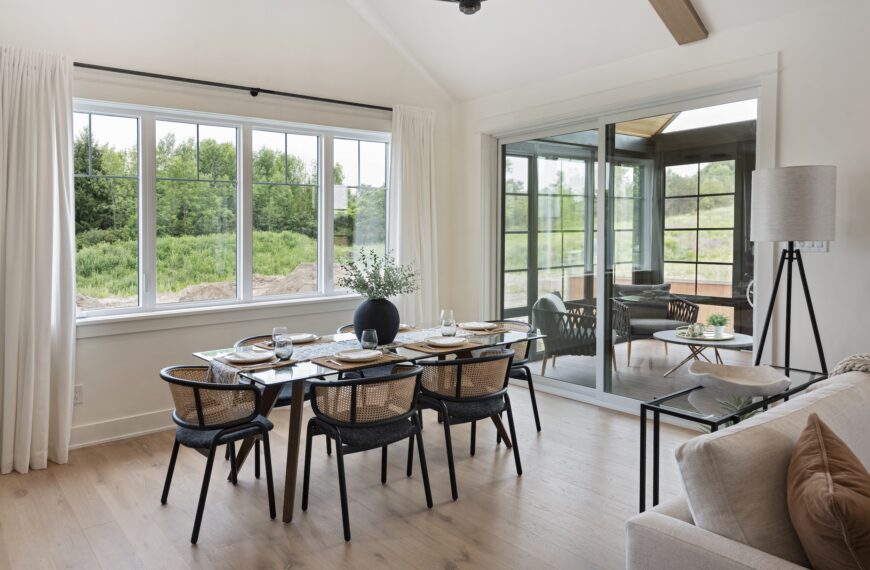 A bright dining area in this Land Ark Homes net zero home features a glass table, six black chairs with cane backs, a sleek vase centerpiece, large windows framing greenery, and glass doors leading to a sunroom with outdoor furniture.