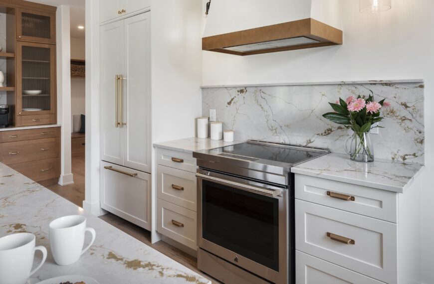 Modern kitchen with white cabinets, marble countertops and backsplash, stainless steel oven, and a range hood with wood accents. A vase of pink flowers sits on the counter—an inviting touch in this Land Ark Homes net zero home.