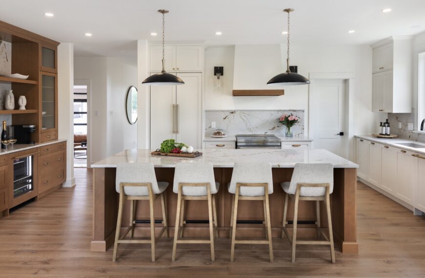 Modern kitchen in a Net Zero Home Building with a large marble island, four upholstered barstools, pendant lights, wooden cabinetry, stainless steel appliances, open shelves, and a vase of flowers near a sunlit window.