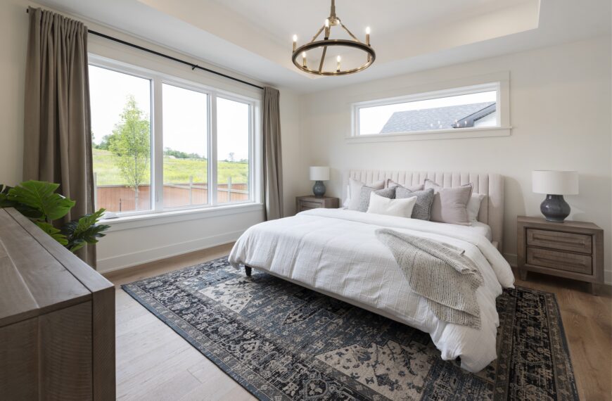 A bright, modern bedroom in a net zero home with a large window, a neatly made bed with white bedding, two wooden nightstands with lamps, a patterned rug, and neutral decor. Natural light fills the room.