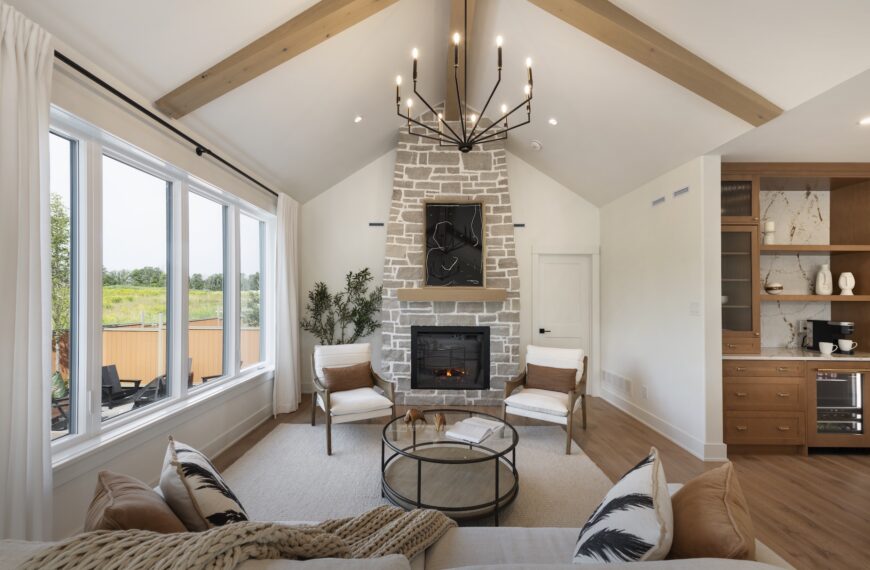 A modern living room with vaulted ceiling, exposed wood beams, large windows, and a stone fireplace showcases the natural light that fills this Production Net Zero Home. Neutral furniture, a round coffee table, and built-in wooden cabinetry complete the inviting space.
