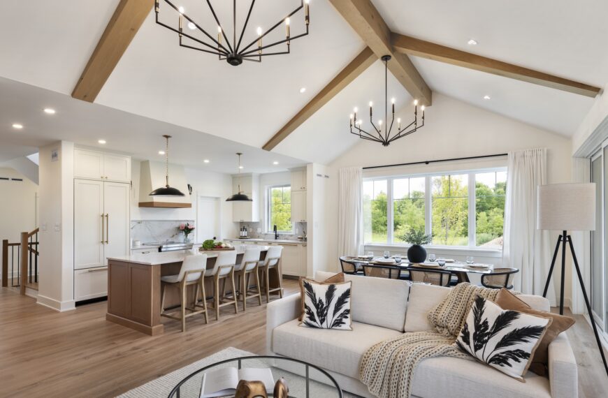 Modern open-concept living area by Land Ark Homes with vaulted ceiling, exposed wooden beams, kitchen island and stools, dining table by large window, neutral-toned sofa with leaf-patterned pillows, and stylish pendant lights.