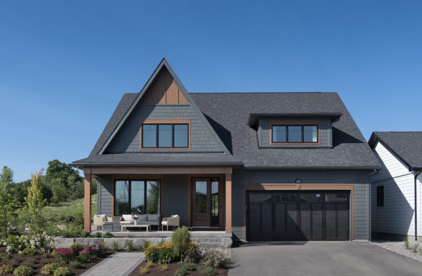 A modern two-story net zero home with dark gray siding, a steep gabled roof, large windows, a double garage, and a front porch with outdoor seating, surrounded by landscaped bushes and plants under a clear blue sky.