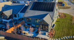 A modern net zero house with solar panels on the roof, a landscaped backyard with stone paths and patio seating, surrounded by a wooden fence in a suburban neighborhood.