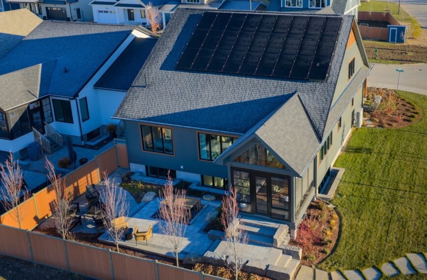 A modern net zero house with solar panels on the roof, a landscaped backyard with stone paths and patio seating, surrounded by a wooden fence in a suburban neighborhood.